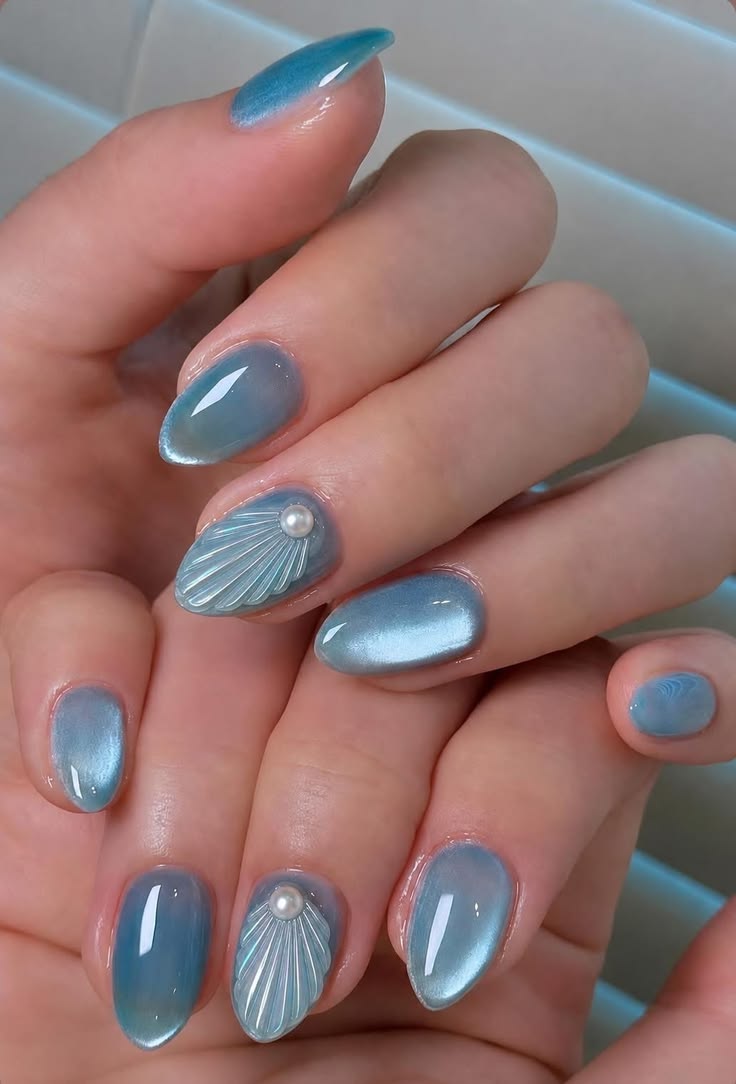 M&atilde;o feminina com unhas ovais pintadas de azul-claro perolado. Os dedos anelares t&ecirc;m uma concha em relevo com uma p&eacute;rola no centro