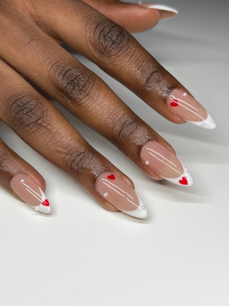 M&atilde;o de uma pessoa com pele escura, unhas longas em formato amendoado, com esmalte nude e pontas brancas, decoradas com cora&ccedil;&otilde;es vermelhos e pequenas p&eacute;rolas brancas
