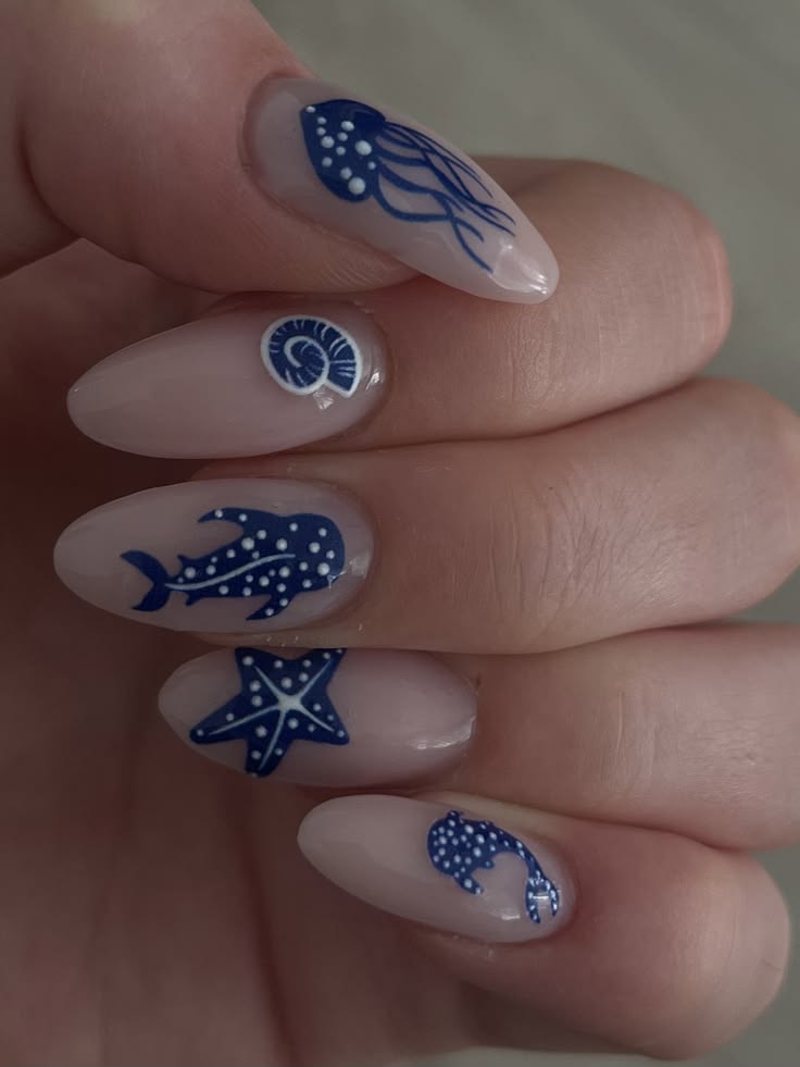 M&atilde;o com unhas ovais em tom nude, cada uma decorada com um adesivo azul marinho de tema marinho: &aacute;gua-viva, concha, tubar&atilde;o-baleia, estrela-do-mar e golfinho