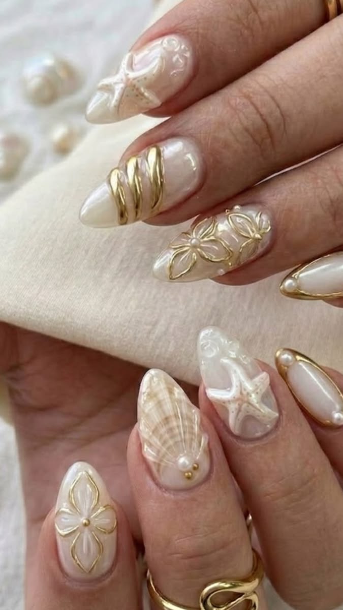 M&atilde;os femininas com unhas longas e pontiagudas, esmalte perolado e decora&ccedil;&otilde;es em relevo dourado e branco, incluindo estrelas-do-mar, conchas, flores e an&eacute;is dourados