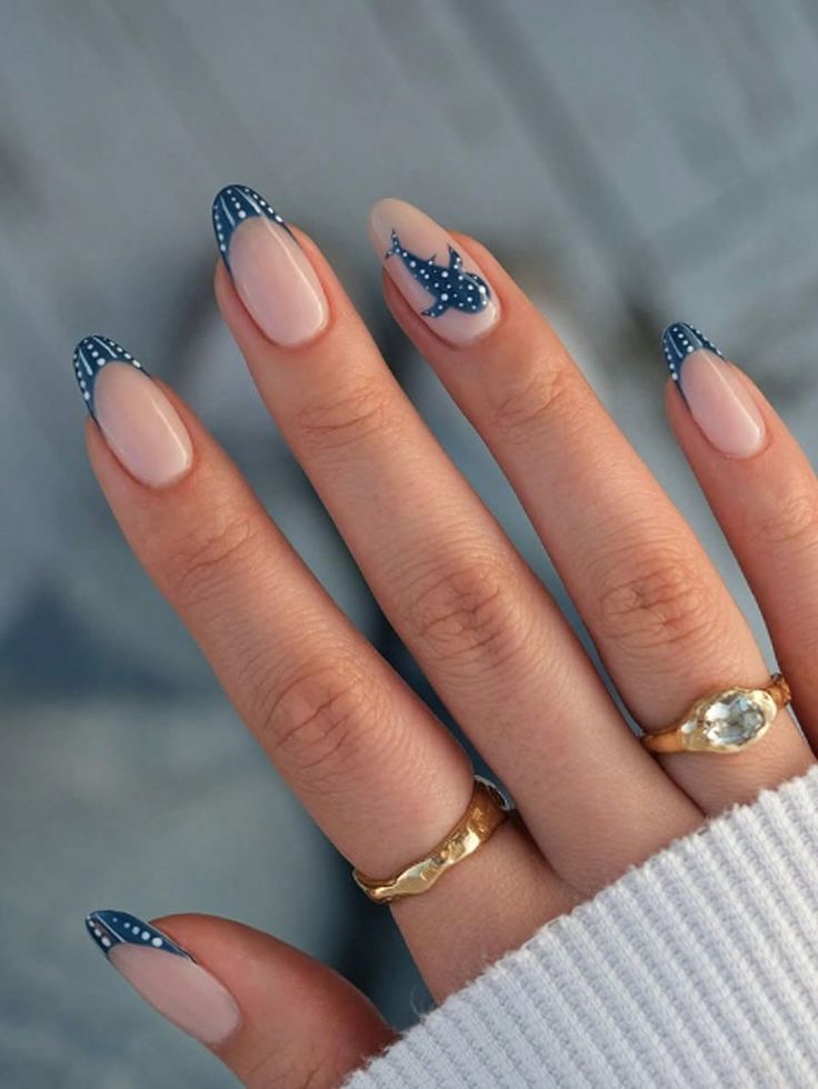 M&atilde;o feminina com unhas ovais e esmalte nude. Tr&ecirc;s unhas t&ecirc;m francesinha azul-marinho com bolinhas brancas, e a unha do dedo anelar tem um tubar&atilde;o-baleia azul-marinho com bolinhas brancas. A m&atilde;o usa dois an&eacute;is dourados e veste uma blusa branca de tric&ocirc;