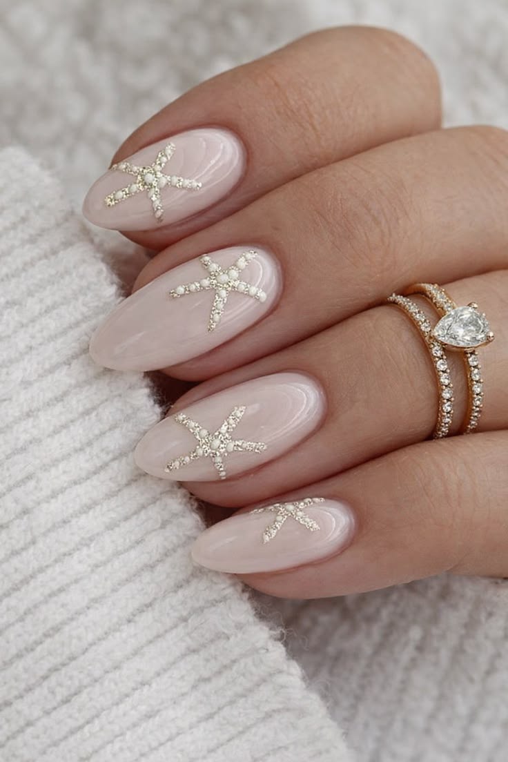M&atilde;o feminina com unhas ovais em tom nude, decoradas com estrelas-do-mar brilhantes em prata e branco. No dedo anelar, um anel duplo dourado com pedras cintilantes e um diamante em formato de gota. A m&atilde;o repousa sobre um tecido de tric&ocirc; branco