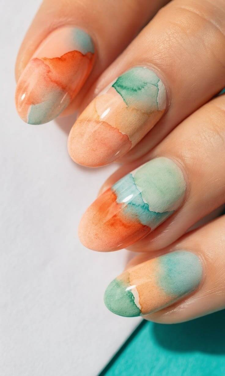 M&atilde;o com unhas ovais pintadas em degrad&ecirc; aquarelado, com tons de laranja, azul-claro e verde-&aacute;gua, sobre fundo branco e azul-turquesa