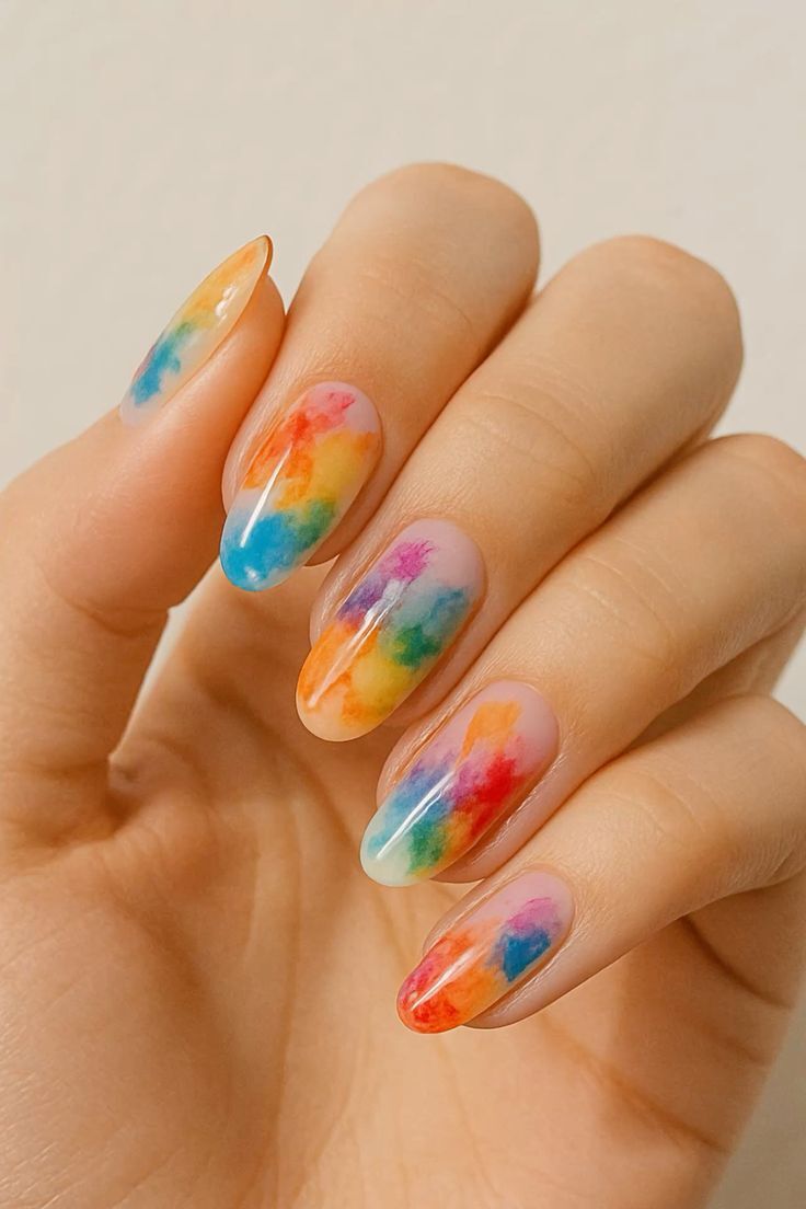 M&atilde;o feminina com unhas longas e ovais, decoradas com esmalte transparente e manchas coloridas em tons de arco-&iacute;ris, como aquarela