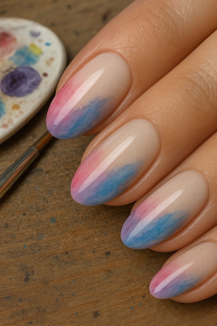 M&atilde;o com unhas ovais decoradas em degrad&ecirc; de rosa, roxo e azul, com base nude. Ao fundo, um pincel e uma paleta de tintas coloridas sobre madeira r&uacute;stica