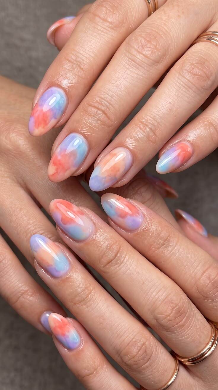 M&atilde;os com unhas ovais pintadas em tons pastel de azul, laranja e roxo, criando um efeito tie-dye. An&eacute;is dourados finos adornam os dedos