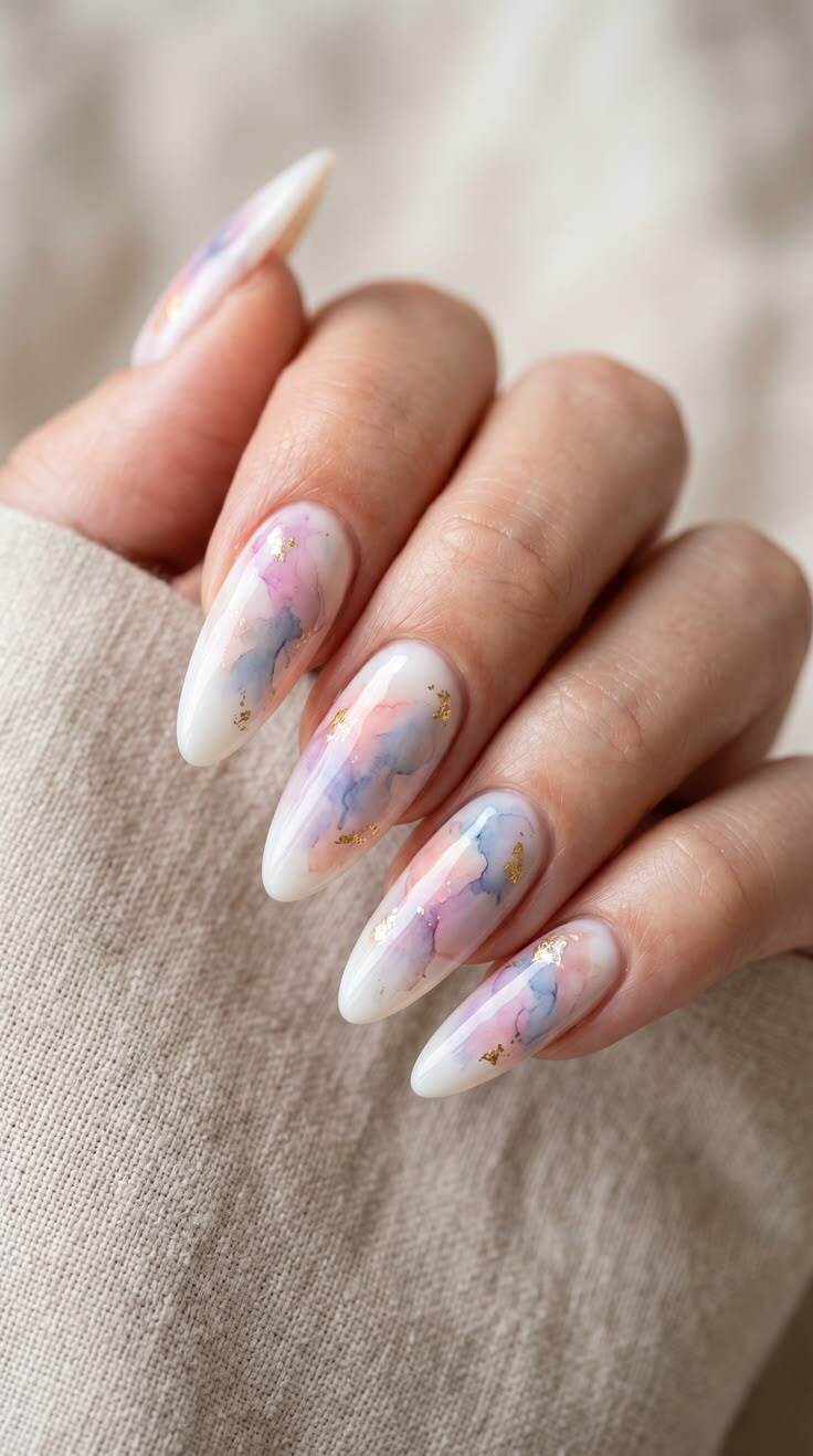 M&atilde;o feminina com unhas amendoadas longas, esmalte branco com manchas abstratas em tons de rosa, azul e roxo, e detalhes de folha de ouro. A m&atilde;o est&aacute; sobre um tecido bege texturizado