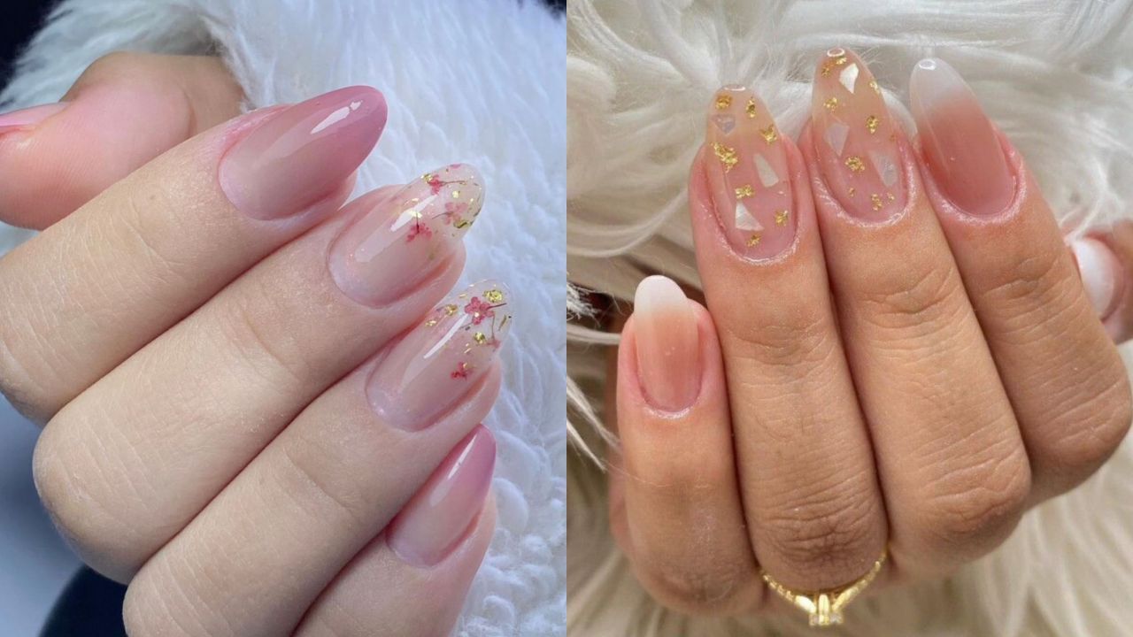 Duas mãos com unhas alongadas em tons de rosa e nude. À esquerda, unhas amendoadas com esmalte rosa translúcido e detalhes de flores secas e folha de ouro. À direita, unhas ovais com esmalte nude, detalhes de madrepérola e folha de ouro, e um anel dourado no dedo anelar. Ambas as mãos estão sobre uma superfície felpuda branca