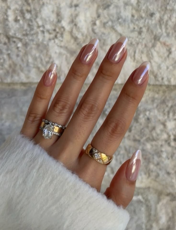 M&atilde;o feminina com unhas amendoadas em esmalte nude perolado e pontas brancas, usando an&eacute;is dourados com diamantes, sobre um fundo de parede clara. A m&atilde;o veste uma manga de su&eacute;ter branco felpudo