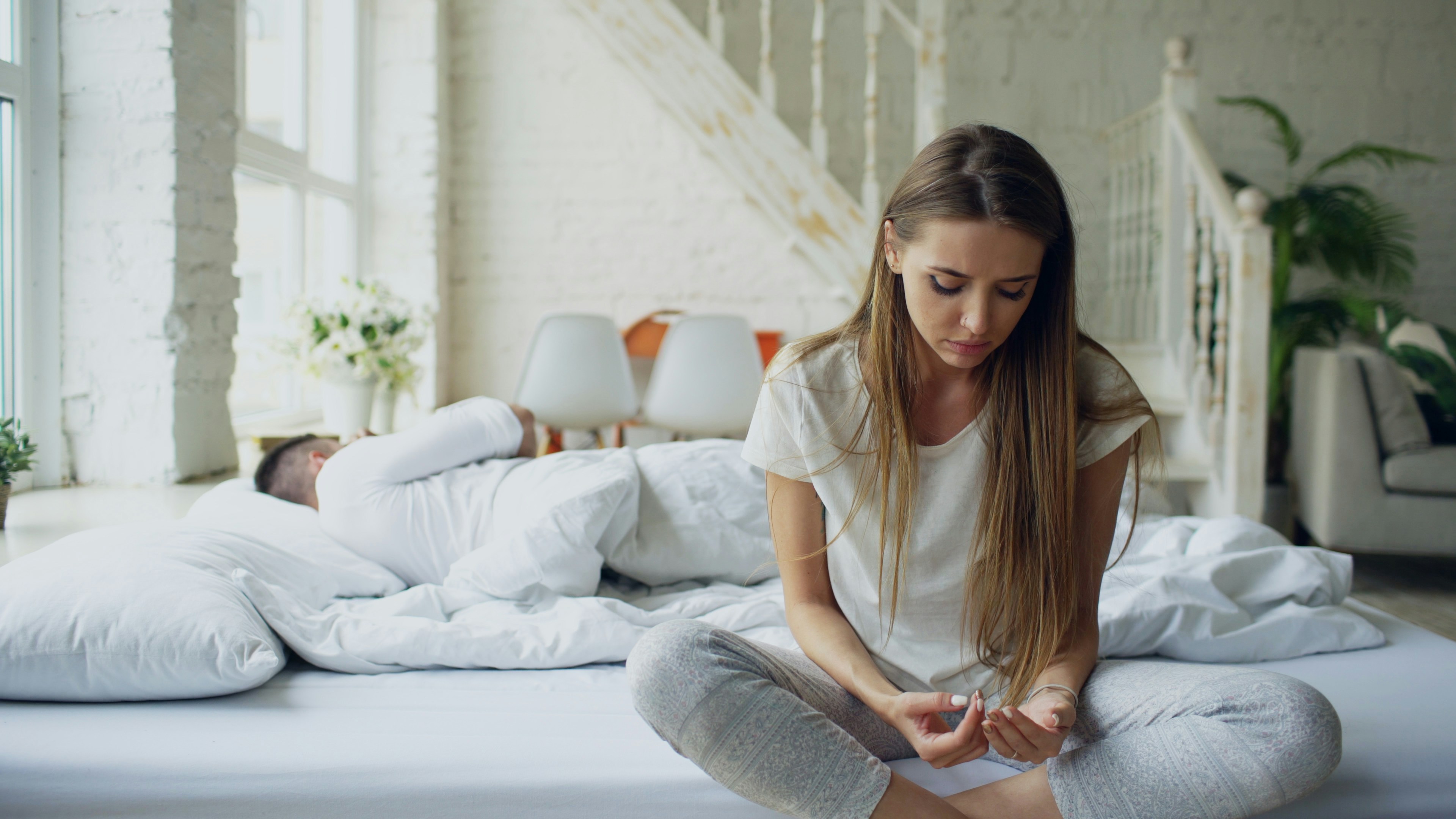 Mulher de cabelos longos e camisa branca sentada na cama, olhando para baixo com express&atilde;o triste, enquanto um homem dorme ao fundo