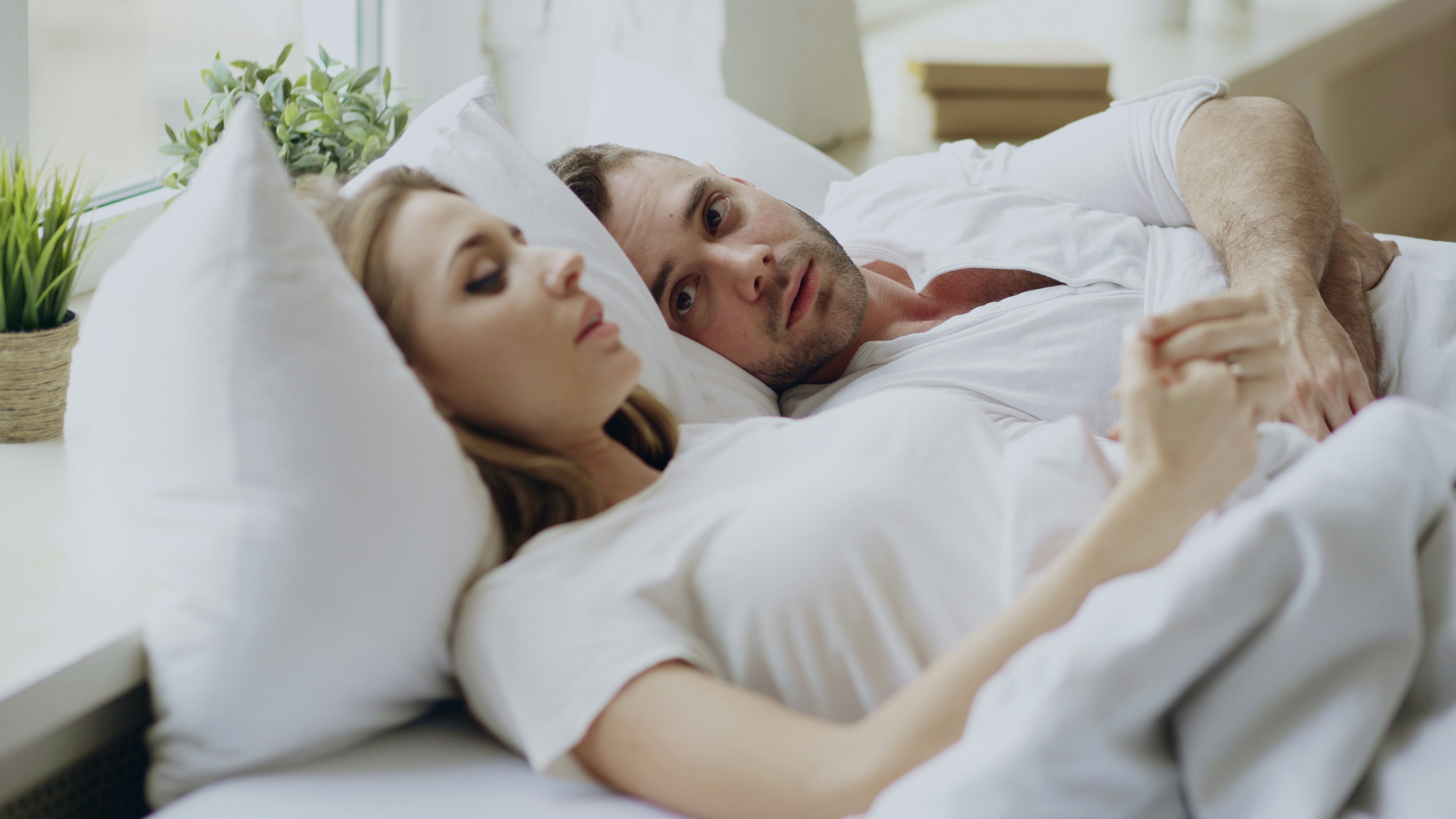Casal deitados na cama, ambos com camisetas brancas. A mulher está deitada de lado, com os olhos fechados, e o homem a observa com expressão séria