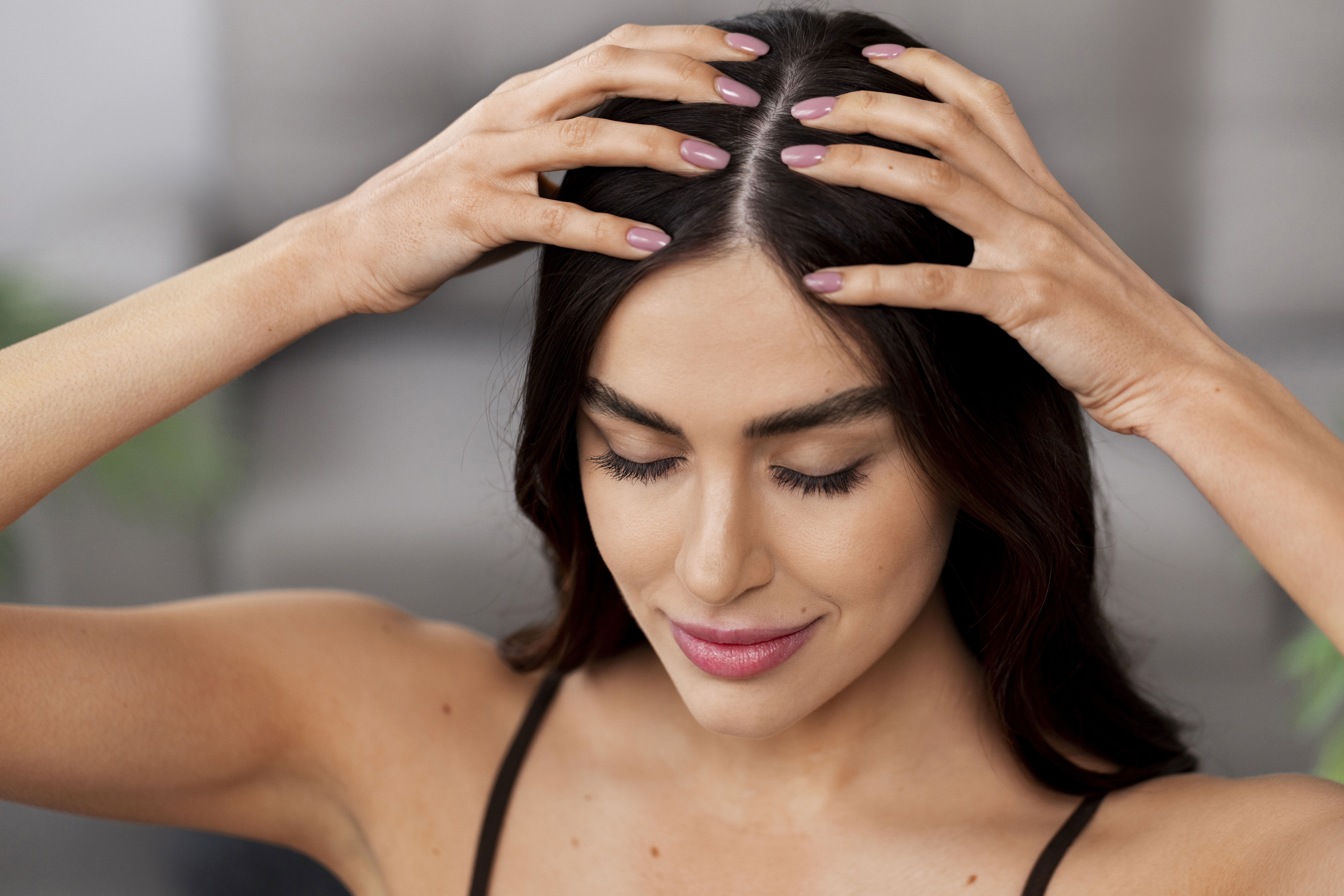 Mulher jovem com cabelos escuros e pele clara massageando o couro cabeludo com as duas m&atilde;os, unhas pintadas de rosa claro. Ela est&aacute; com os olhos fechados e um leve sorriso, vestindo uma al&ccedil;a fina preta, em um fundo desfocado e claro