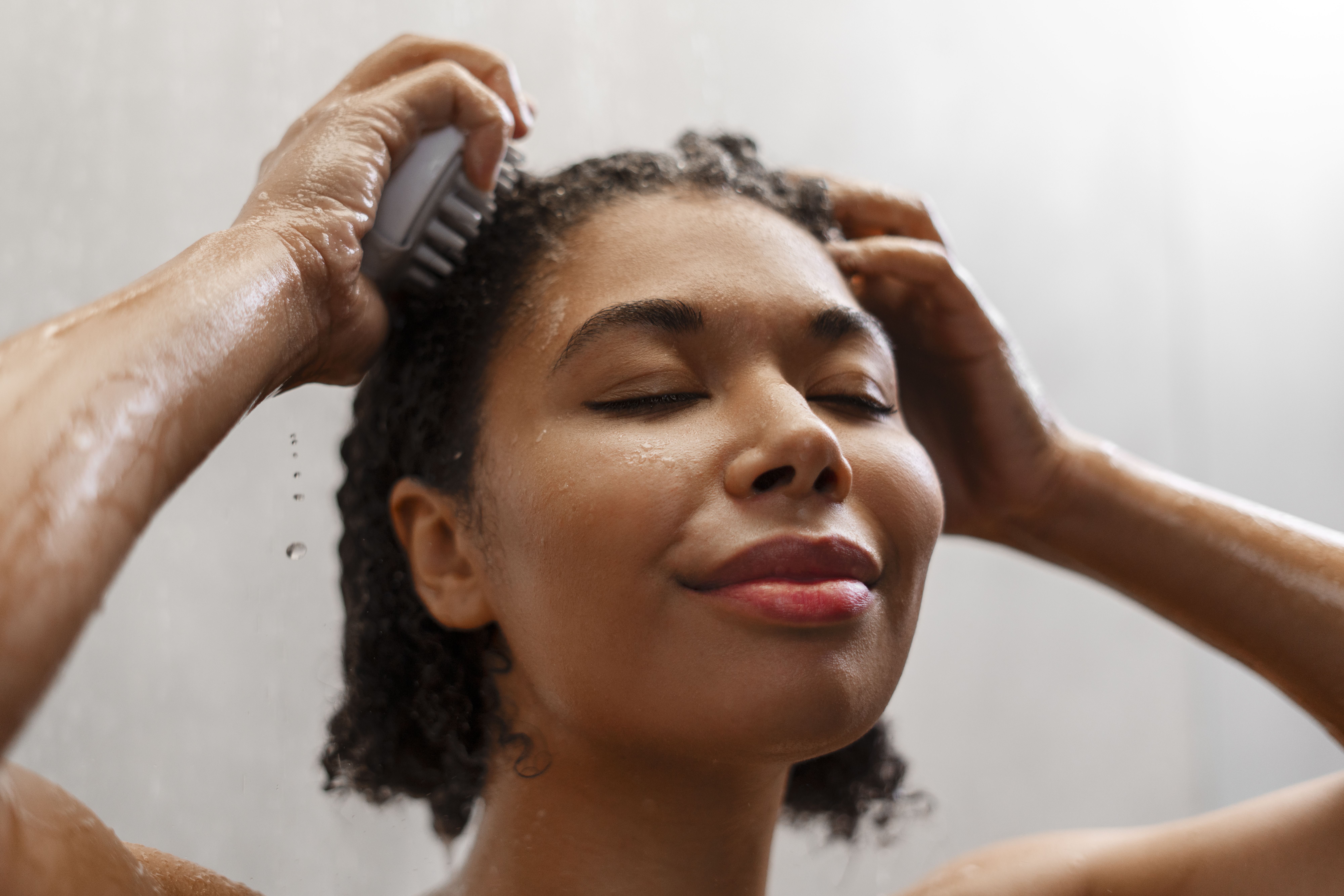 Mulher de pele morena e cabelos crespos lavando a cabe&ccedil;a no chuveiro, usando uma escova massageadora. Ela tem os olhos fechados e um sorriso suave, com gotas de &aacute;gua escorrendo pelo rosto e bra&ccedil;os