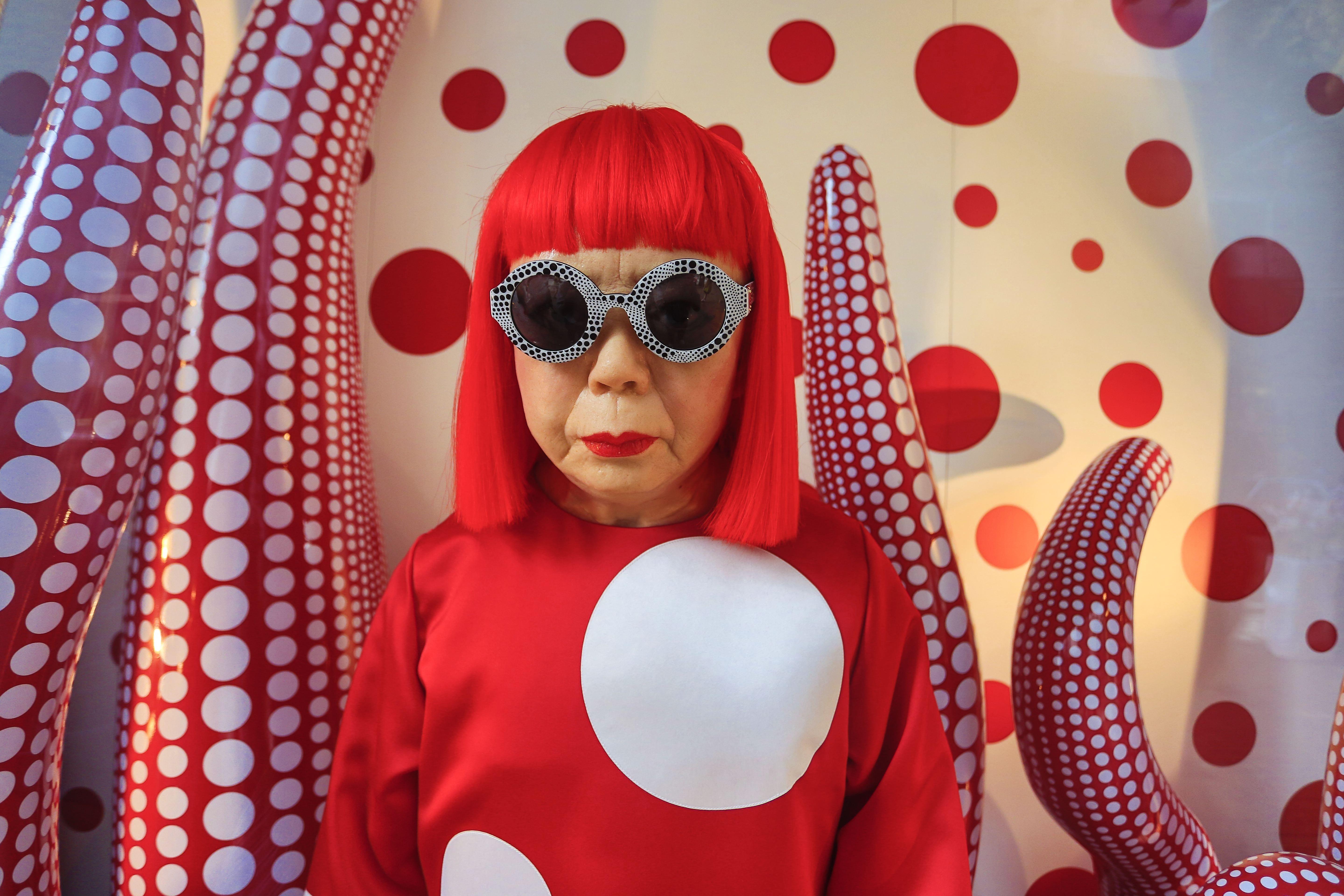Manequim da artista Yayoi Kusama com peruca vermelha, óculos de sol pretos com bolinhas brancas e batom vermelho, vestindo blusa vermelha com bolinhas brancas, em meio a esculturas vermelhas com bolinhas brancas