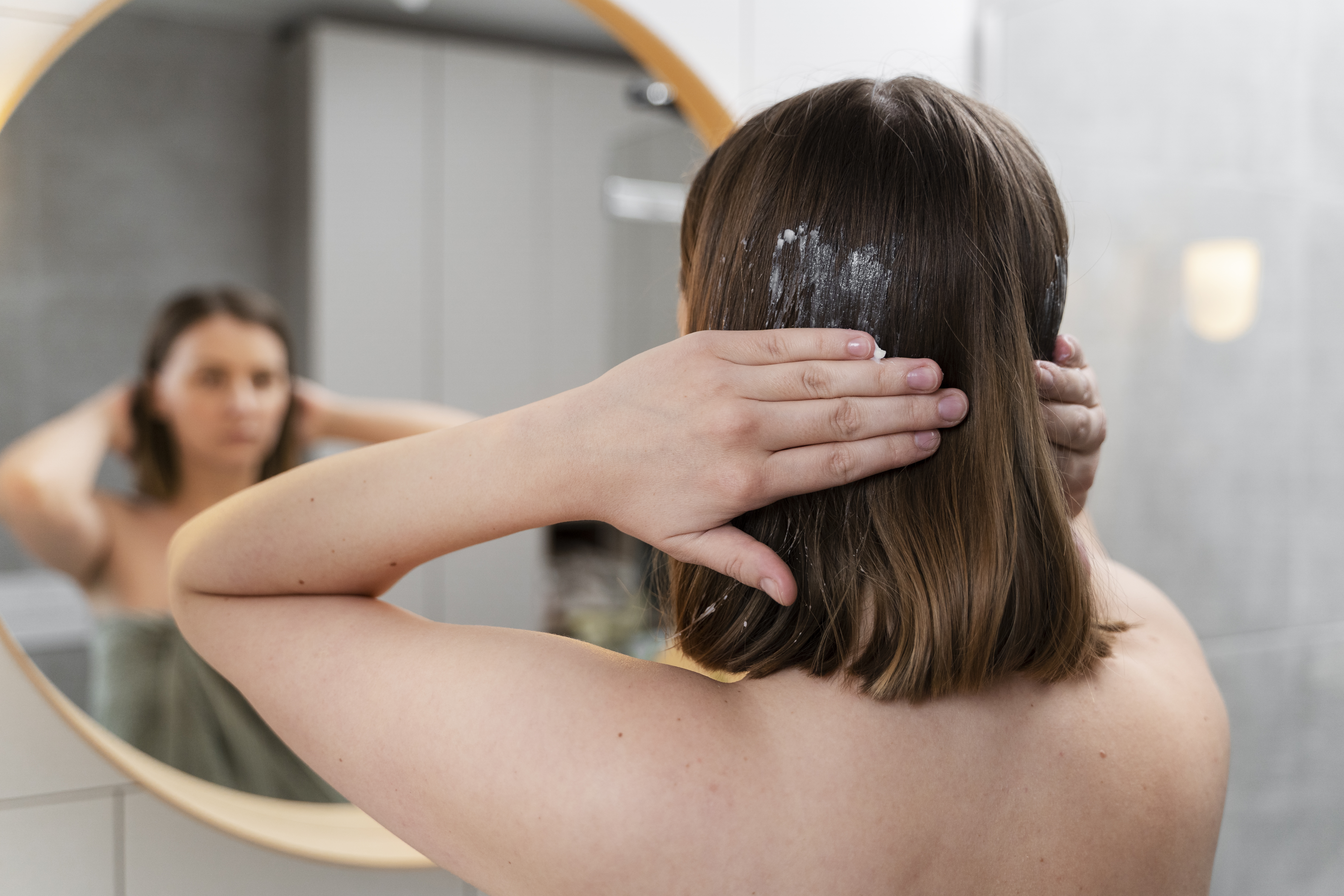 Mulher de costas, com cabelos castanhos curtos, aplicando creme branco no cabelo. Ela se olha no espelho redondo de moldura clara, onde sua imagem aparece emba&ccedil;ada, vestindo uma toalha verde. O fundo &eacute; um banheiro com azulejos brancos e cinzas