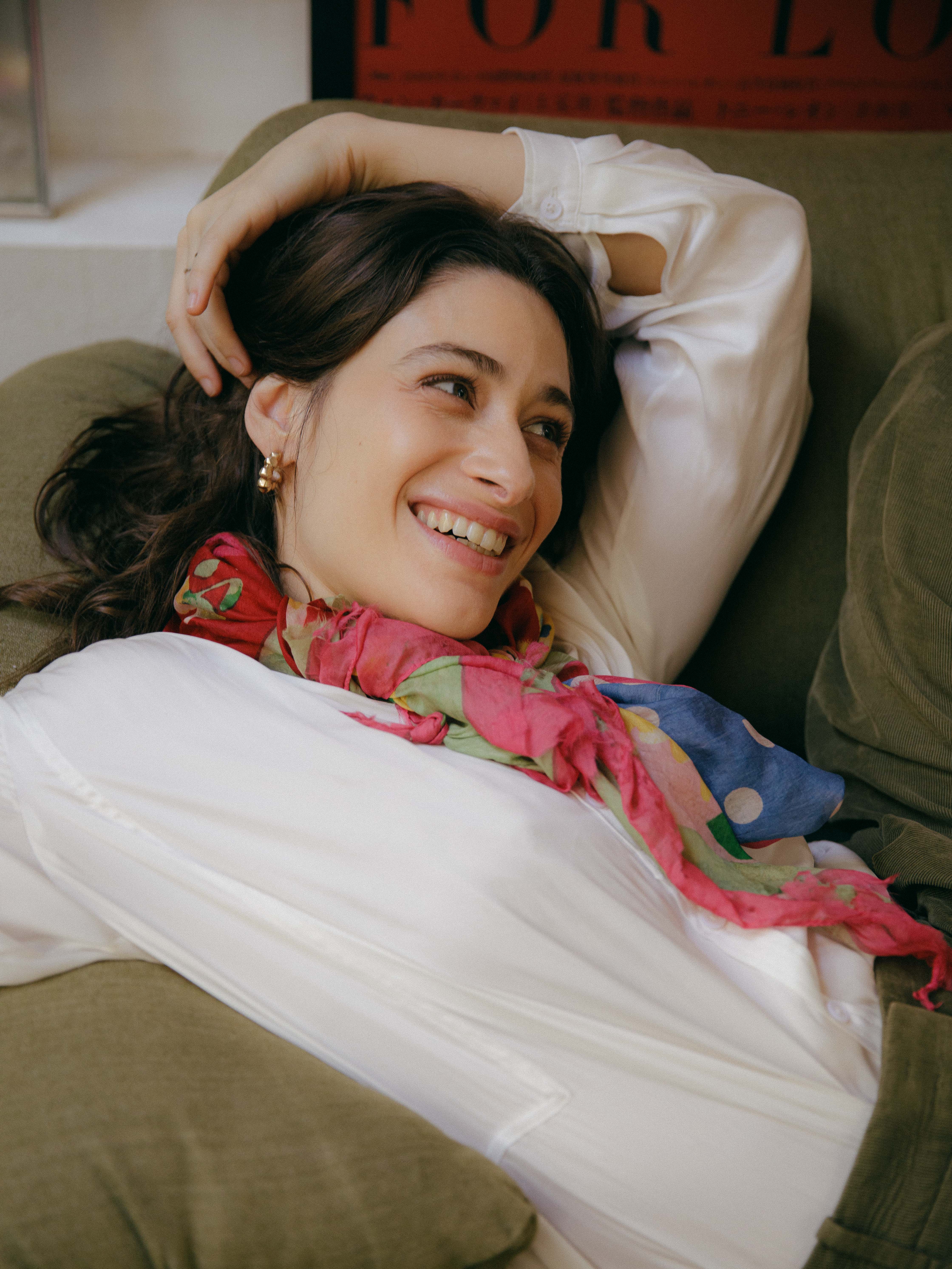 Mulher jovem de cabelos castanhos escuros, sorrindo e olhando para o lado, deitada em um sof&aacute; verde-oliva. Ela veste uma camisa branca e um len&ccedil;o colorido com tons de rosa, verde e azul, e usa brincos dourados. Um bra&ccedil;o est&aacute; atr&aacute;s da cabe&ccedil;a