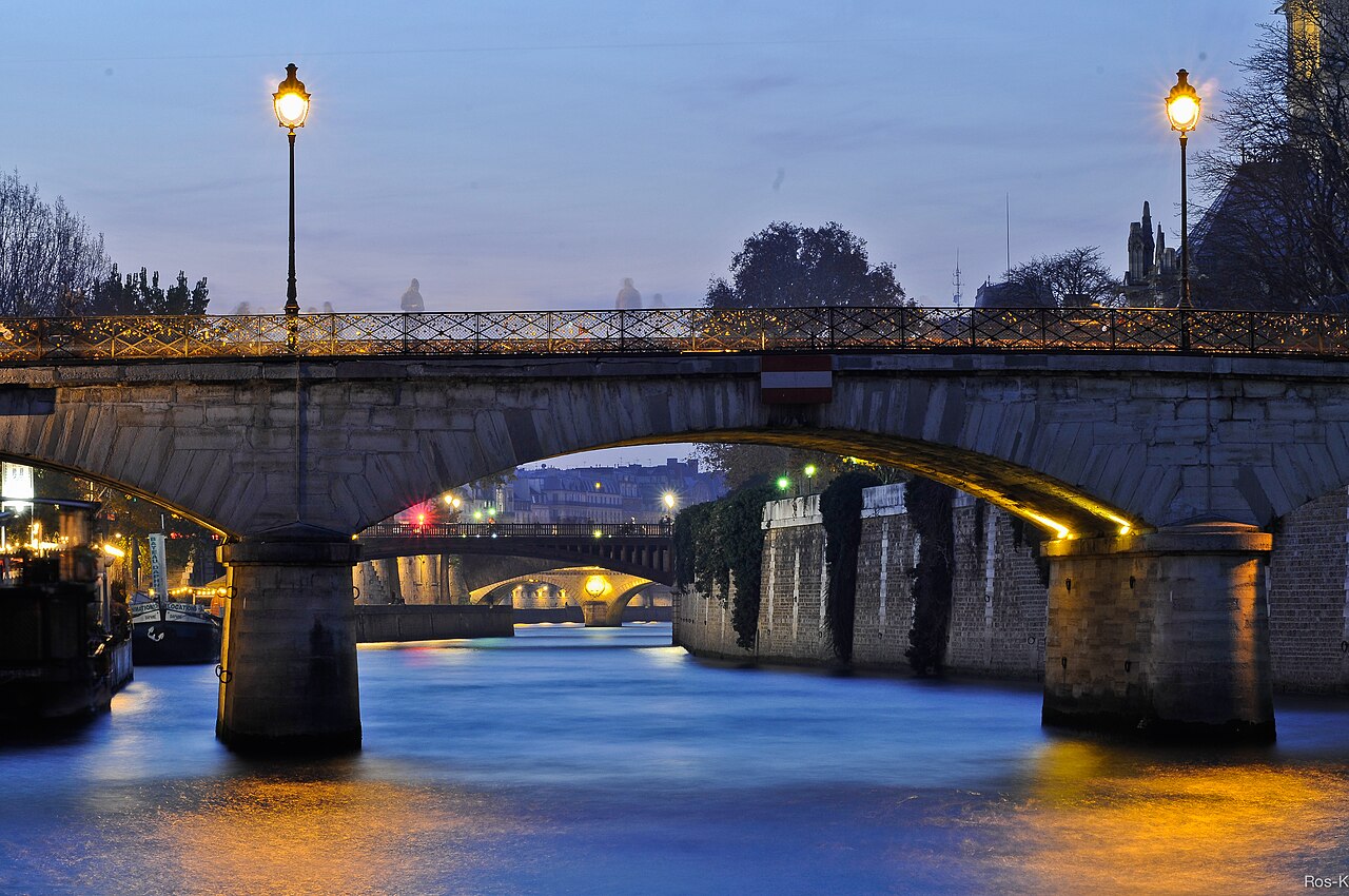 Ponte de pedra iluminada sobre um rio azul-escuro ao anoitecer, com postes de luz acesos e reflexos dourados na água. Outras pontes e edifícios distantes também estão iluminados, sob um céu crepuscular