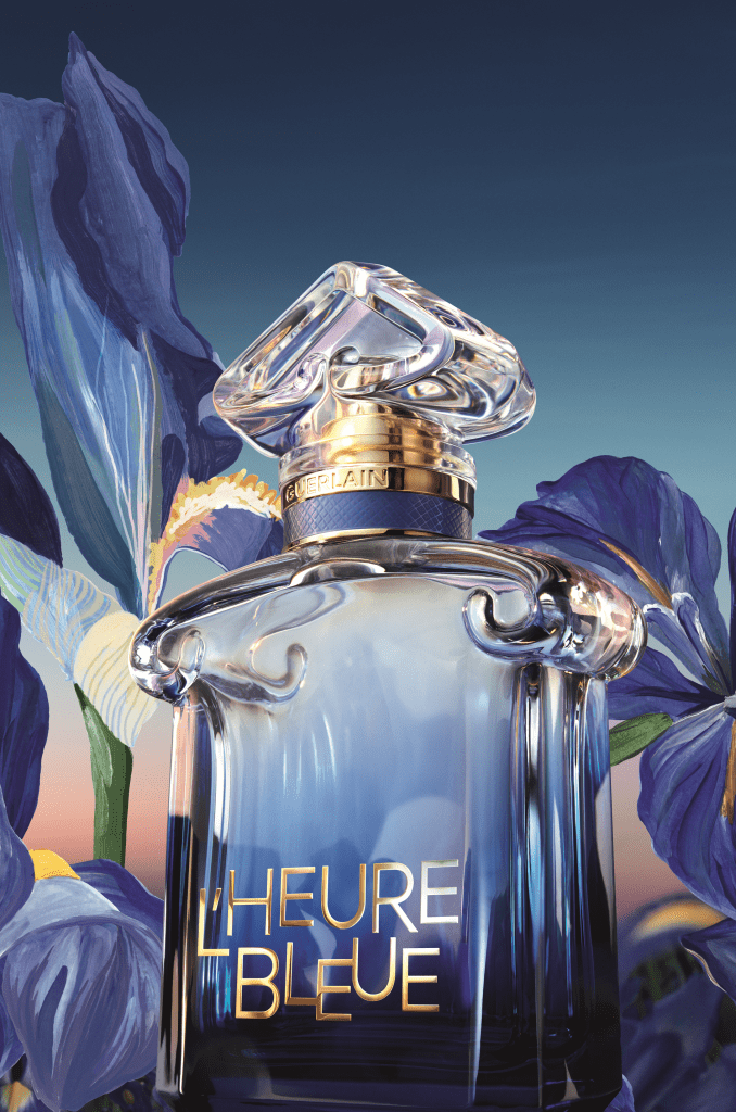 Frasco de perfume Guerlain LHeure Bleue em vidro azul degrad&ecirc; com tampa transparente e detalhes dourados, cercado por flores de &iacute;ris azuis e roxas em um fundo azul claro