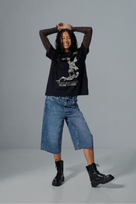 Jovem mulher sorridente com cabelos escuros e cacheados, vestindo camiseta preta com estampa de esqueleto, blusa de manga comprida transparente por baixo, bermuda jeans larga e coturnos pretos, posando com as m&atilde;os na cabe&ccedil;a em fundo cinza