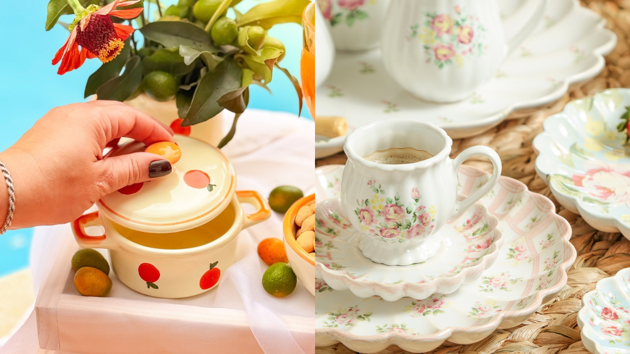 Mão feminina com pulseira prateada e unhas escuras segurando a tampa de uma panela decorada com maçãs vermelhas. Ao lado, xícaras e pratos de porcelana com estampa floral rosa, sobre uma toalha de mesa e um fundo azul.