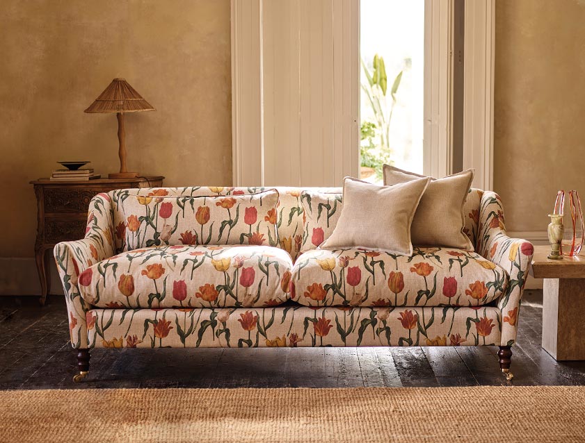 Sof&aacute; de dois lugares com estampa floral de tulipas vermelhas e amarelas, com duas almofadas bege, em uma sala com paredes claras e piso de madeira escura. &Agrave; esquerda, uma mesa de cabeceira de madeira entalhada com um abajur de palha e livros. Ao fundo, uma porta aberta revela um jardim com plantas verdes